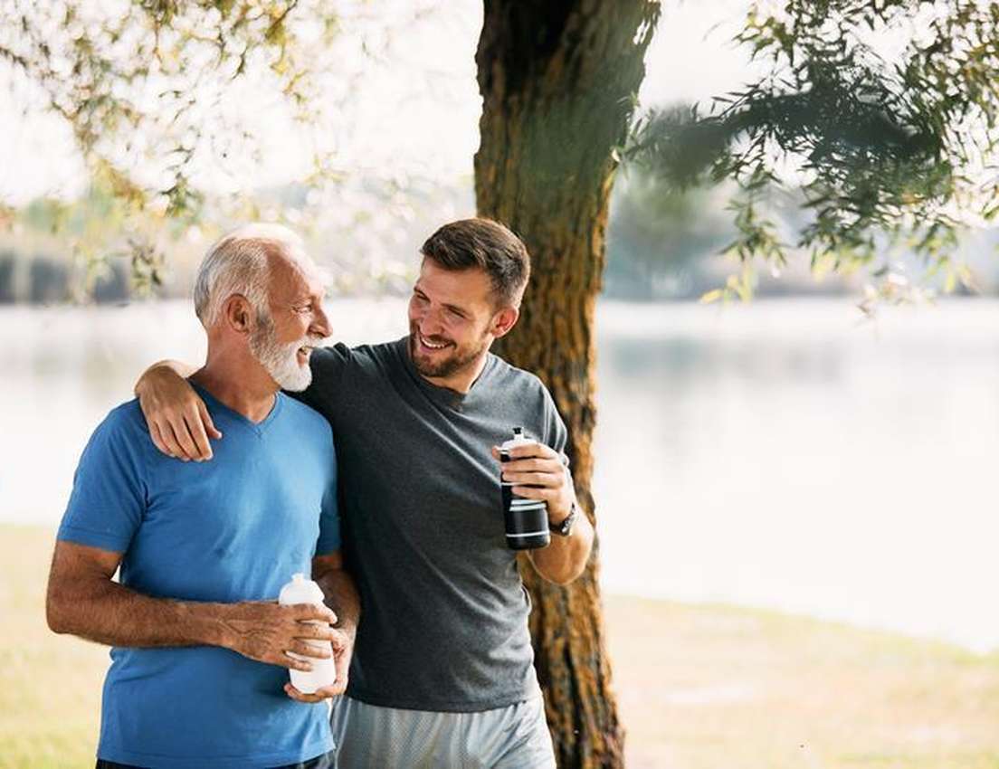 Two men smiling and talking by a lakeside, holding water bottles. One has his arm around the other's shoulder, with a tree in