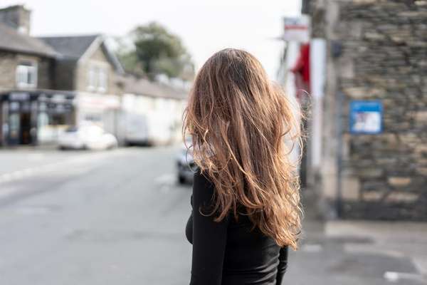 A person with long, wavy brown hair, seen from behind, stands on a street with blurred stone buildings and parked cars.