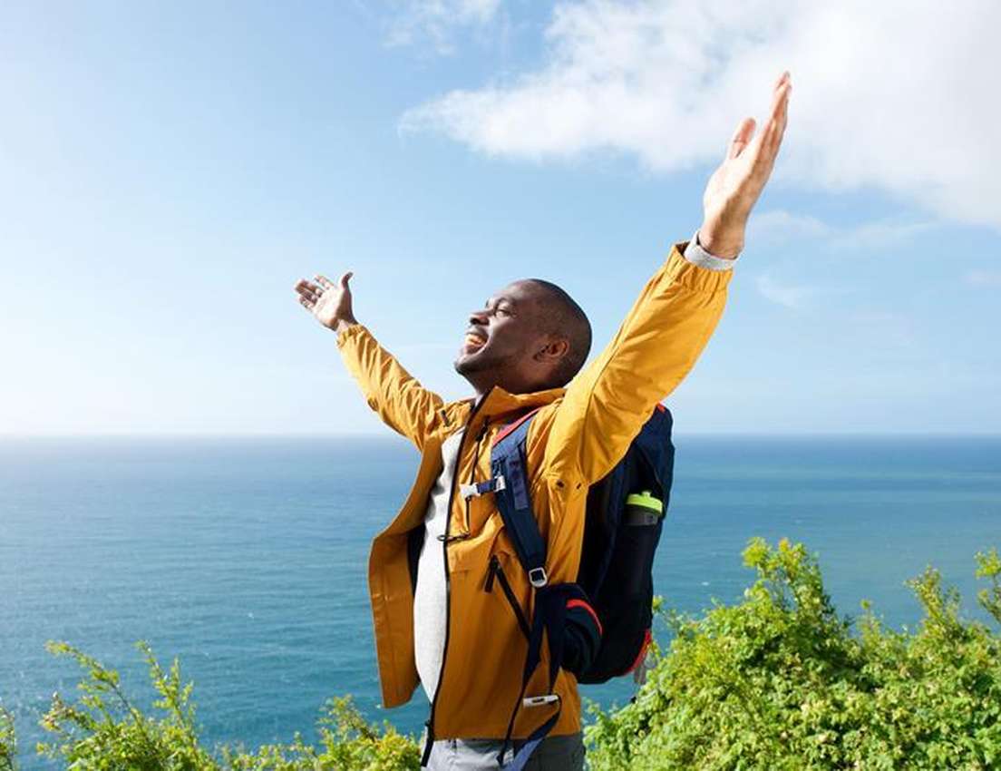 Man in yellow jacket with backpack joyfully raises arms by the ocean, standing on a green cliff under a clear blue sky.