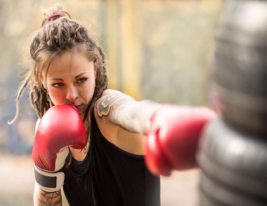 Woman with tattoos wearing red boxing gloves punches a hanging bag outdoors, focusing intently.
