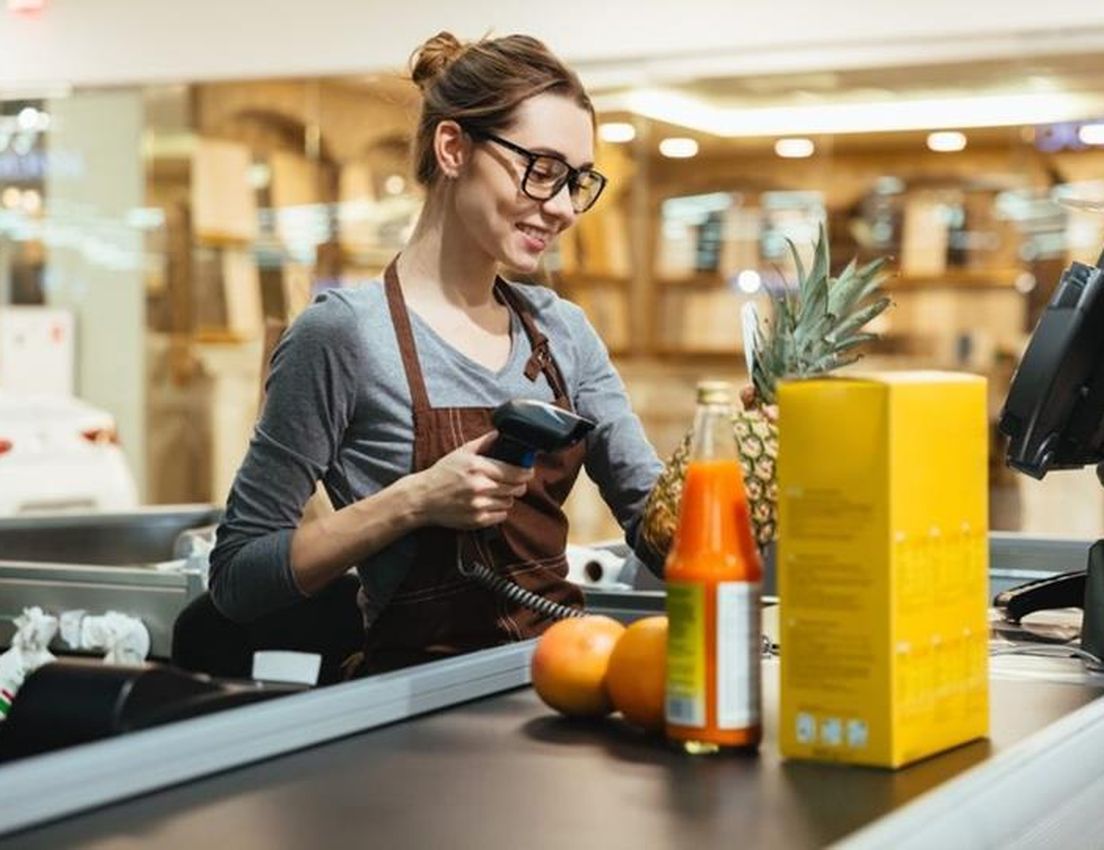 Cashier with glasses scanning groceries, including a pineapple, orange juice, and oranges, at a supermarket checkout counter.