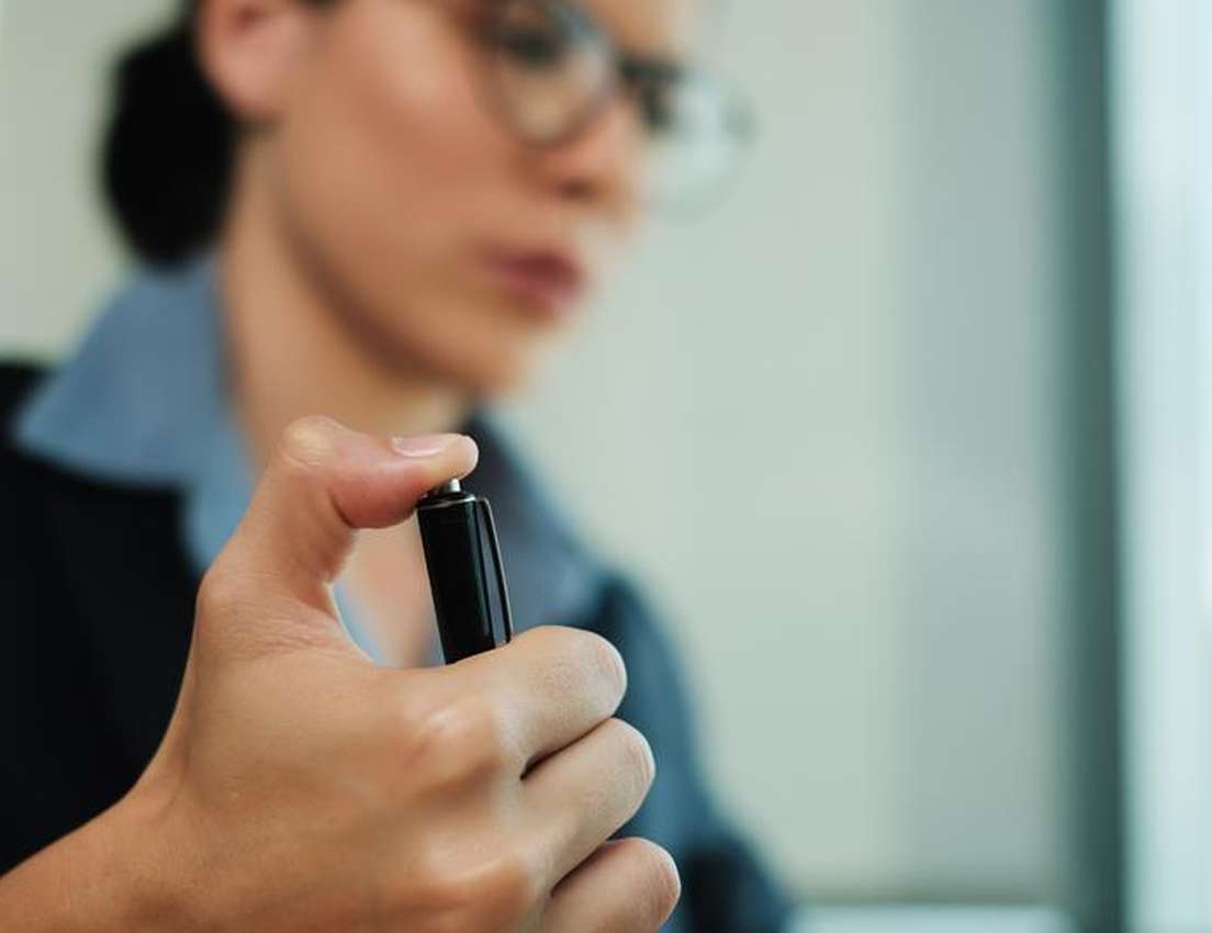 Close-up of a person in glasses and a blazer holding a black pen with a focused expression, set in an office environment.