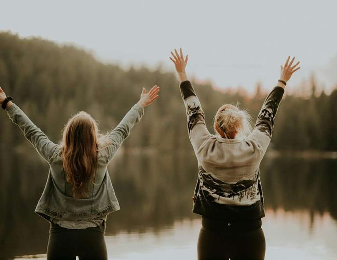 Two people raising their arms in joy, facing a serene lake with a forested backdrop at sunset.