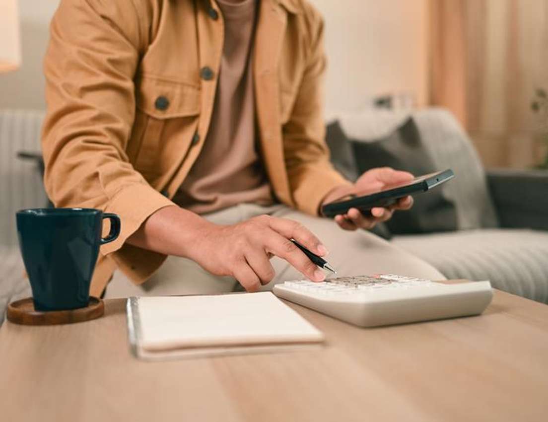 Person in brown jacket calculates on a white calculator, holding a smartphone, with a mug and notepad on a wooden table.