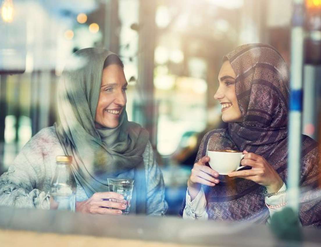 Two smiling women in hijabs chat in a cafe. One has coffee, the other water. Seen through a window with light reflections.