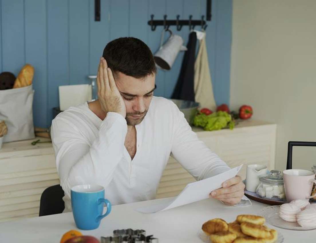 Stressed man with hand on head reads a document at a kitchen table with a blue mug and pastries.