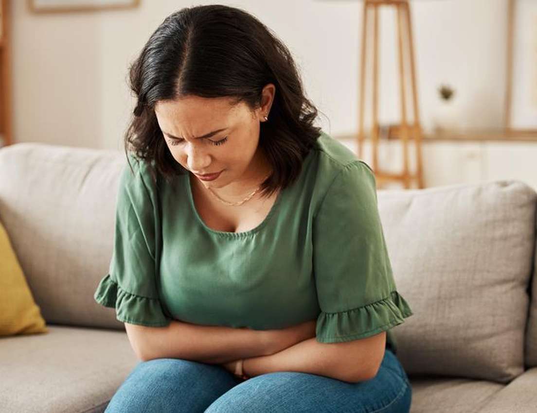 A woman on a couch clutches her stomach with a pained expression, suffering from abdominal pain.