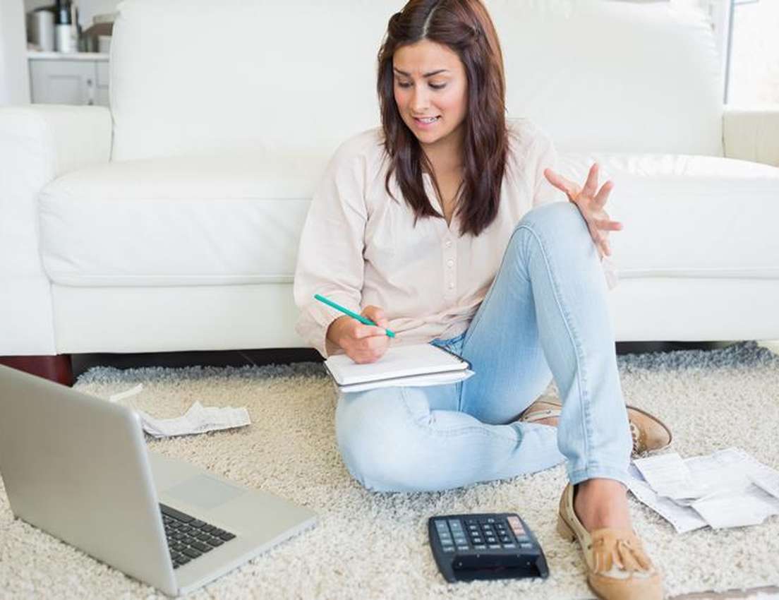 A frustrated woman sits on a rug, surrounded by a laptop, calculator, and papers, writing in a notebook, managing finances.