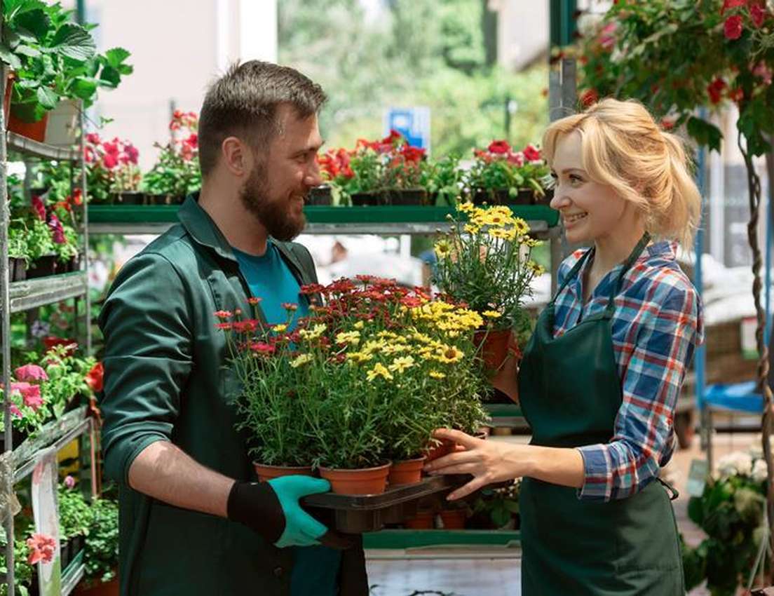 Smiling man and woman exchanging trays of potted red and yellow flowers at a garden center.