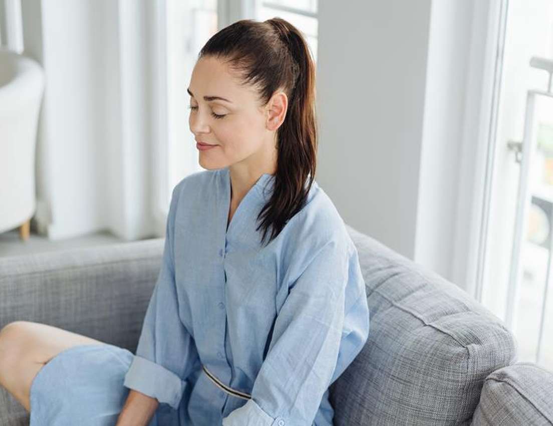 A woman in a blue dress sits peacefully on a gray couch with eyes closed, enjoying a relaxed moment in a bright, airy room.
