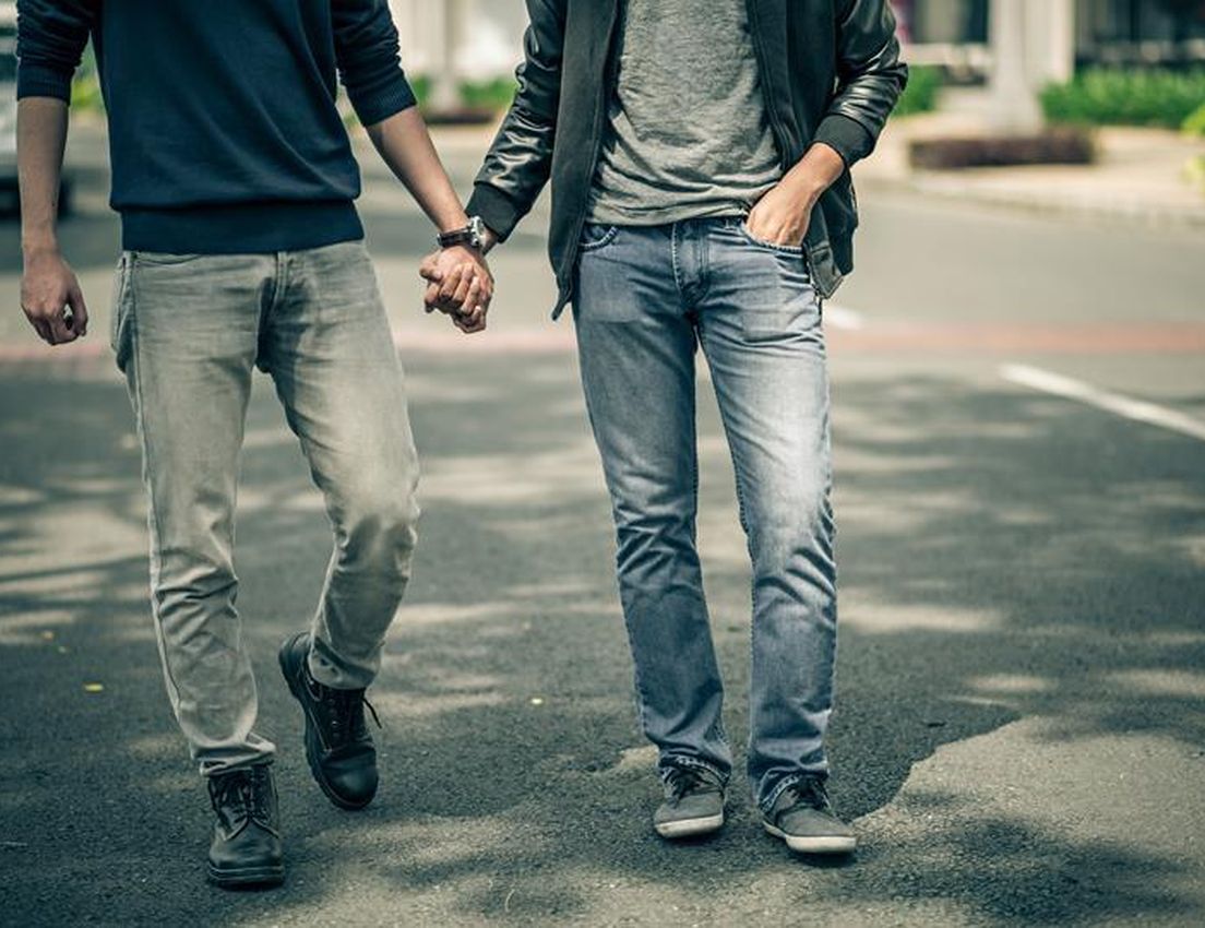 Two people in jeans and casual tops walk hand-in-hand on a paved surface. One person has a hand in their pocket.