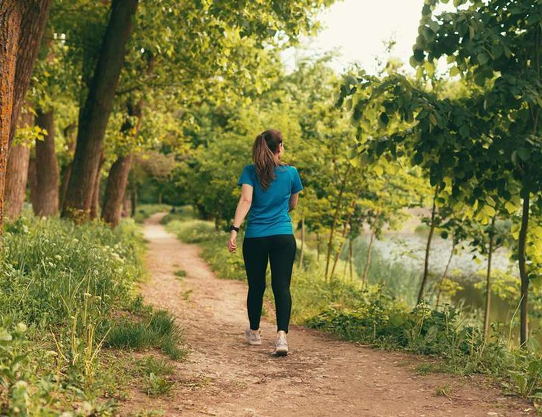 From behind, a woman in a blue shirt and black leggings walks on a winding dirt path through lush trees beside a lake.