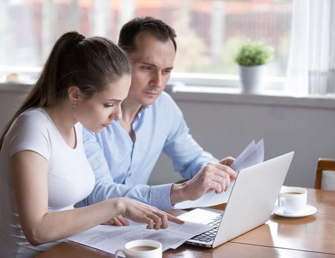A man and woman sit at a table, intently reviewing documents and a laptop. The woman points at papers. Coffee cups are nearby