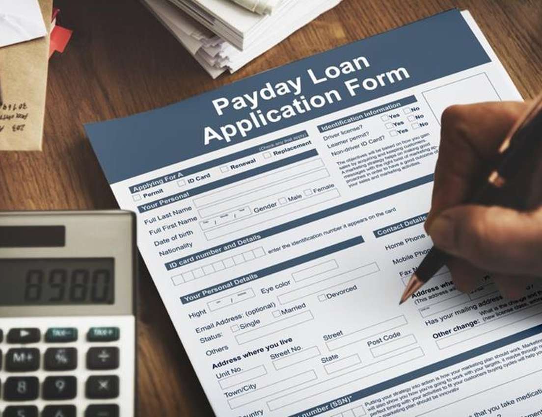 A hand fills out a Payday Loan Application Form on a wooden desk, with a calculator showing "8980" and stacked papers nearby.