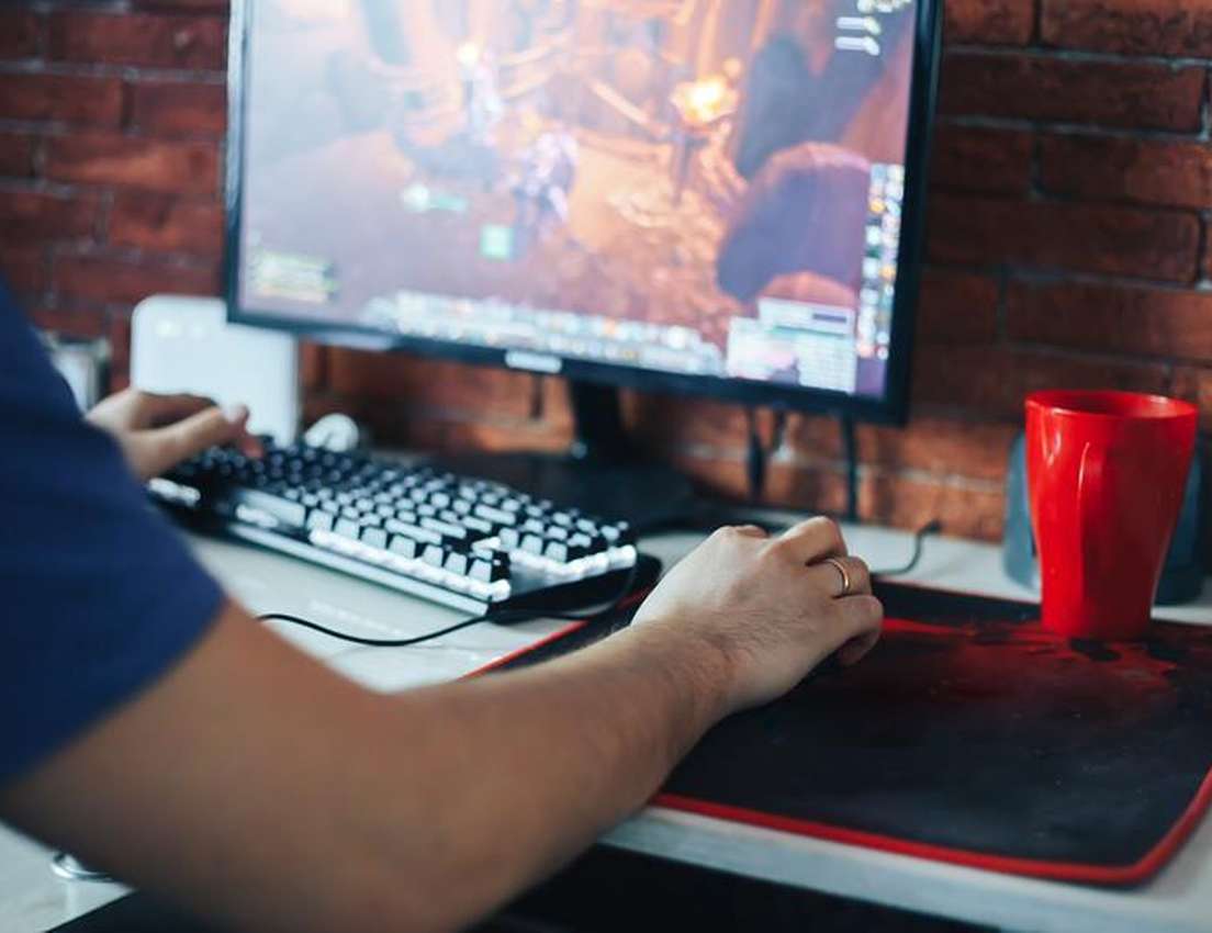 Person's hands using a mechanical keyboard and mouse to play a video game on a computer monitor. A red cup sits on a black mo