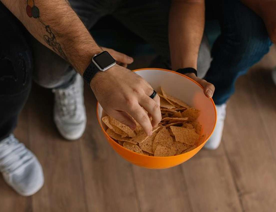 Overhead shot of a person with a smartwatch and arm tattoo holding an orange bowl filled with tortilla chips.