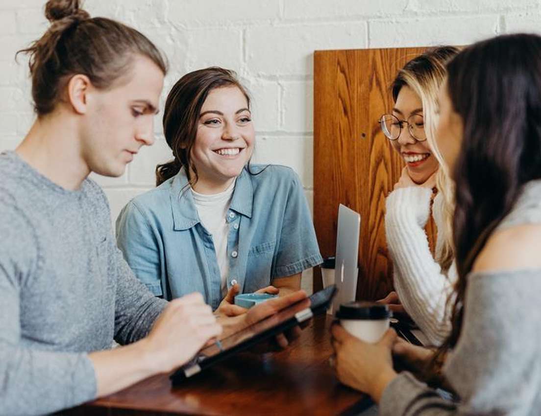 Four diverse young adults smiling and interacting at a table with coffee and a tablet, in a cafe-like setting.