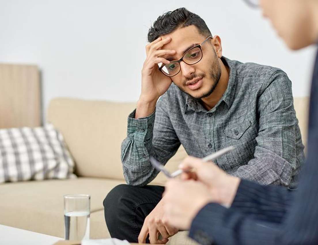 Man in glasses holds head, talking to another person with notepad and pen. They are seated on a beige sofa with a glass of wa