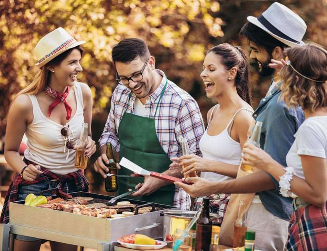 A group of friends happily barbecuing in a park, enjoying drinks and laughter, with one person wearing a green apron tending 