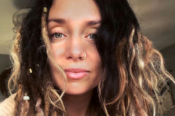 Close-up of a woman with blue eyes, a sparkly nose ring, and dark, wavy hair with blonde streaks and dreadlocks adorned with