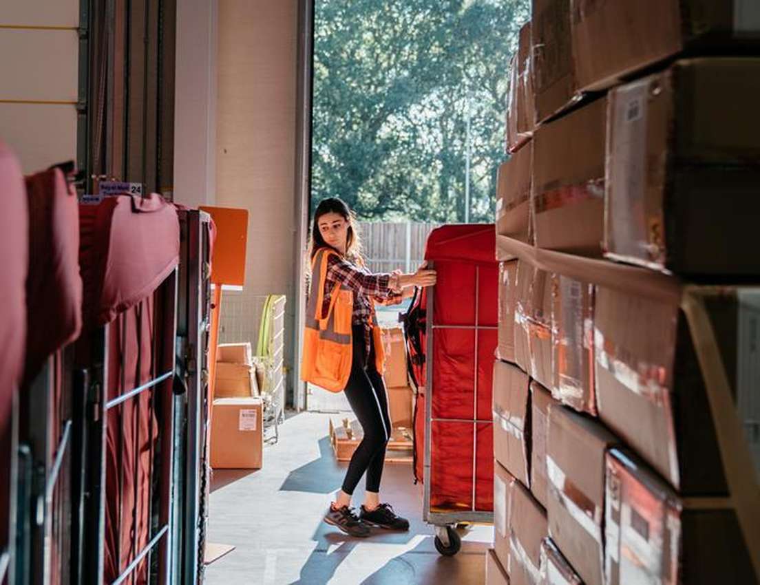 A young woman in an orange safety vest and plaid shirt pushes a red mail cart through a sunny warehouse loading bay, surround