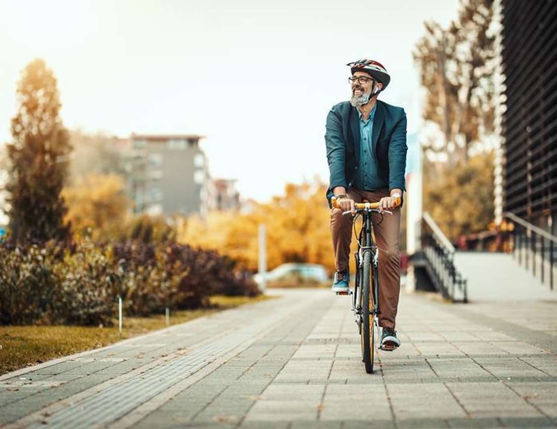Smiling bearded man in a helmet and jacket rides a bicycle on a sunny urban path lined with autumn trees.
