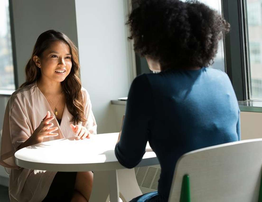 Two women conversing at a white table by a sunny window. One smiles and gestures while facing forward; the other is seen from