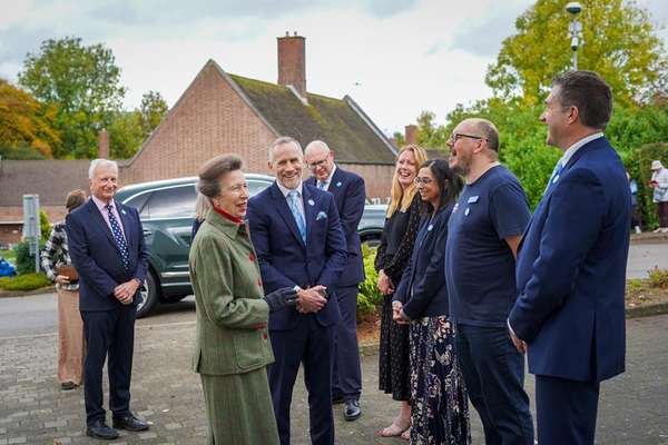 Princess Anne, in a green tweed suit, laughs with a diverse group of staff and officials wearing blue pins at an outdoor even