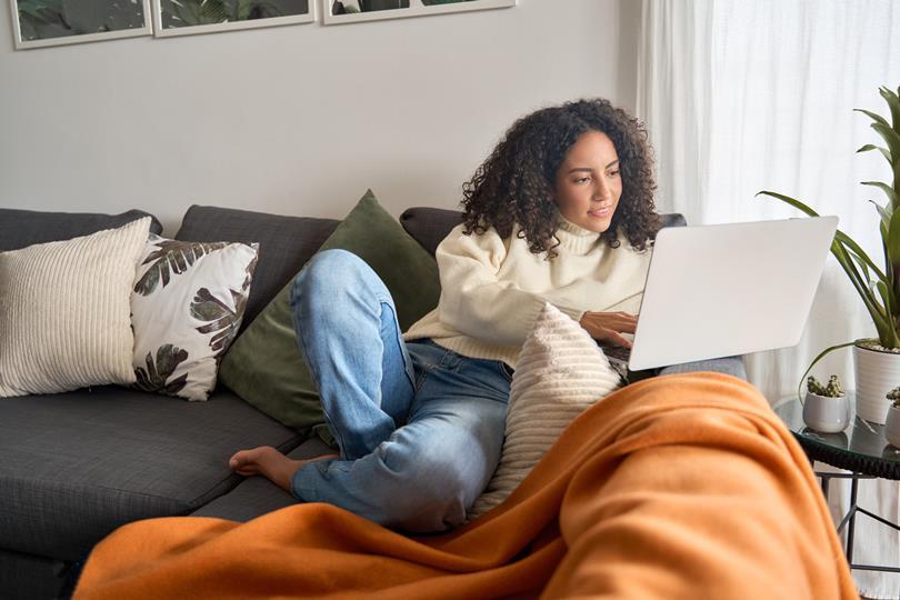 A young woman with curly hair in a cream sweater and jeans relaxes comfortably on a gray couch with pillows, using a laptop. An orange blanket covers her legs.