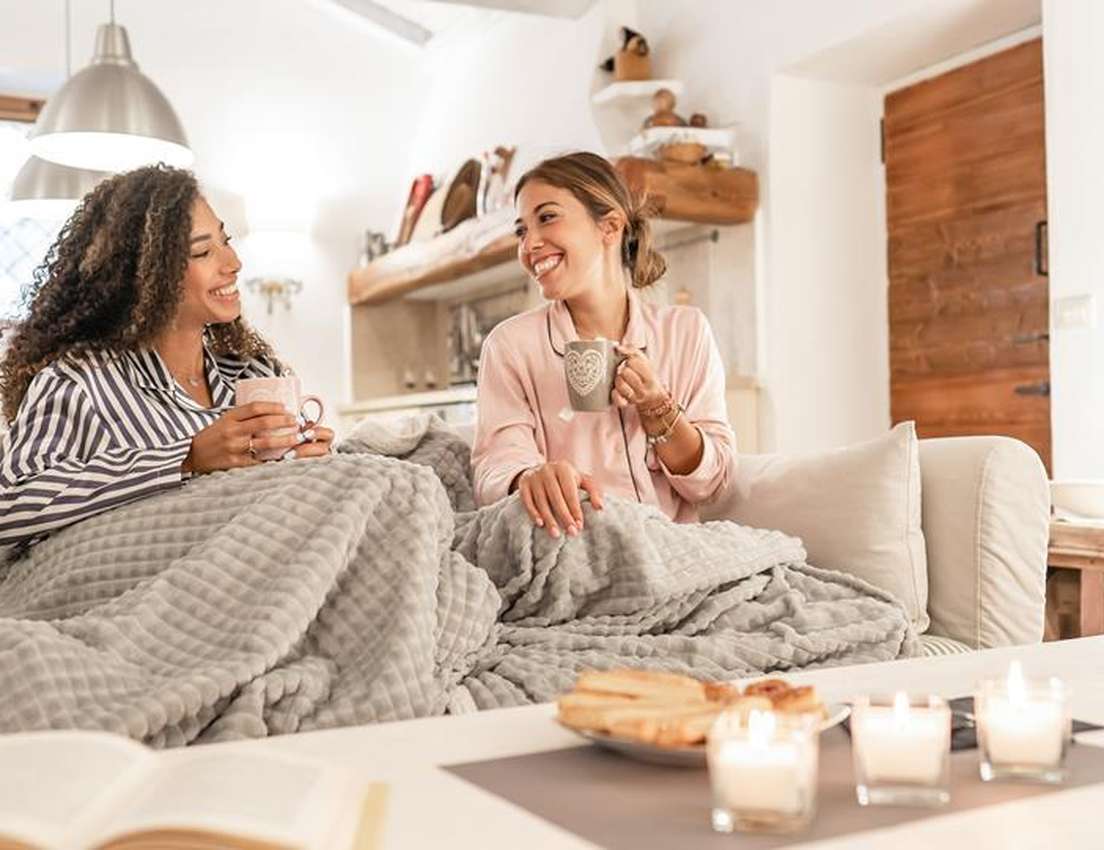Two smiling women in pajamas on a couch under a blanket, drinking from mugs. Candles and pastries are on a table in the foreg