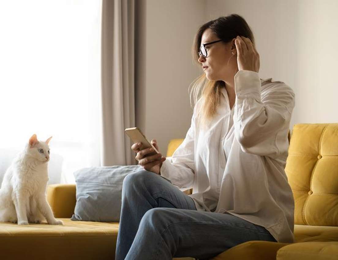 A woman with glasses on a yellow couch holds a phone and touches her ear, next to a white cat looking out a bright window.