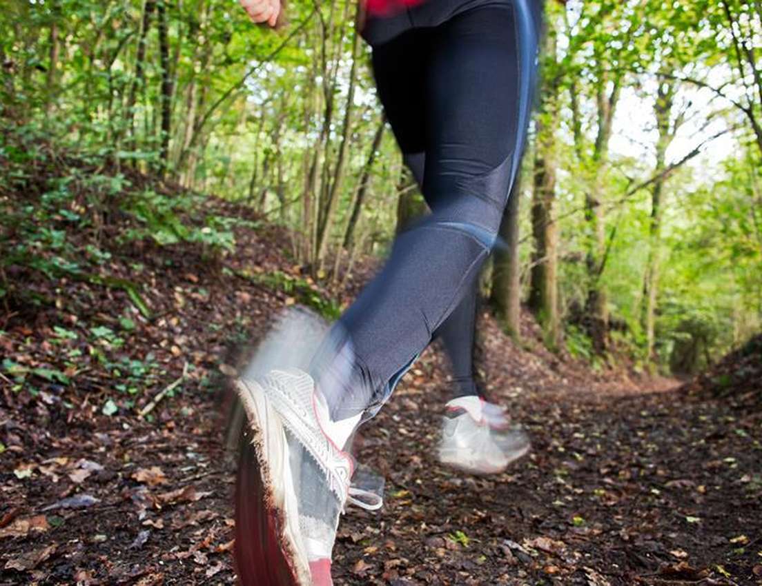 A person's legs in black leggings and sneakers run fast on a leaf-strewn forest trail, blurred by motion.