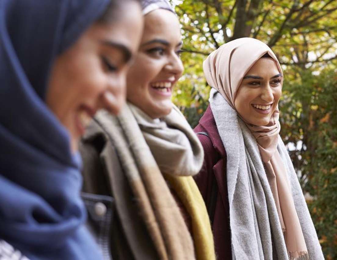 Three young women in hijabs laugh happily outdoors. The woman on the right, in a pink hijab and grey scarf, smiles brightly.
