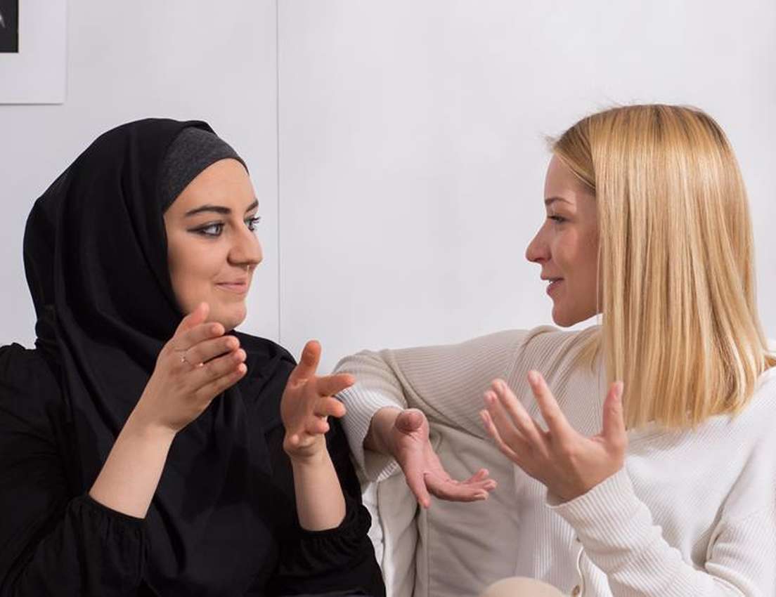 Two women sitting on a sofa, engaged in an animated conversation; one wears a hijab, the other has blonde hair.