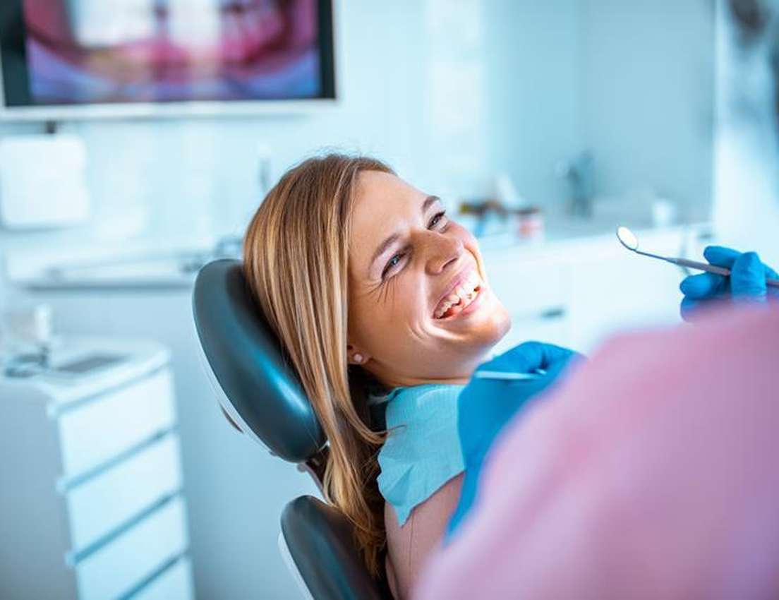 Beaming woman in dental chair laughs up at dentist's gloved hand with mirror. Bright dental office setting.
