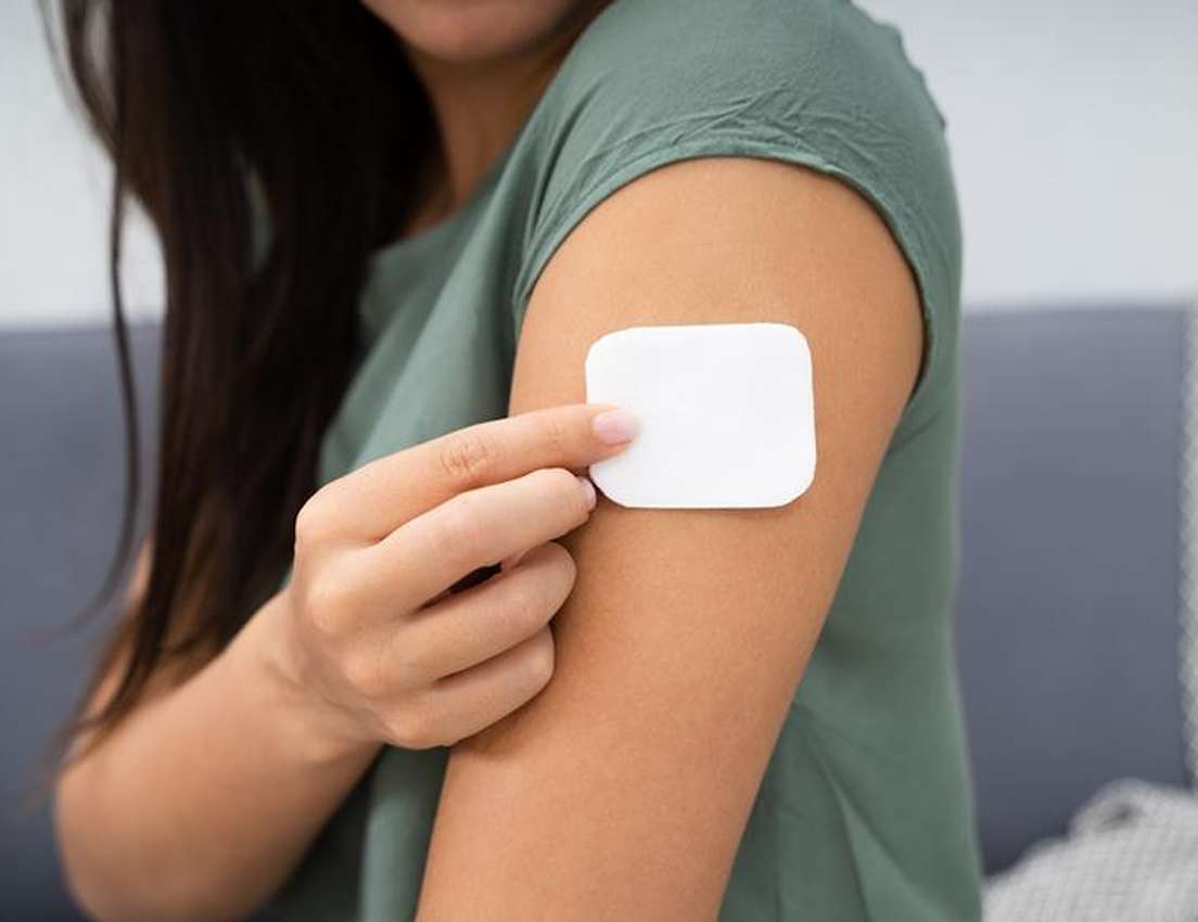 A person with long dark hair and a green shirt touches a white square patch applied to their upper arm.