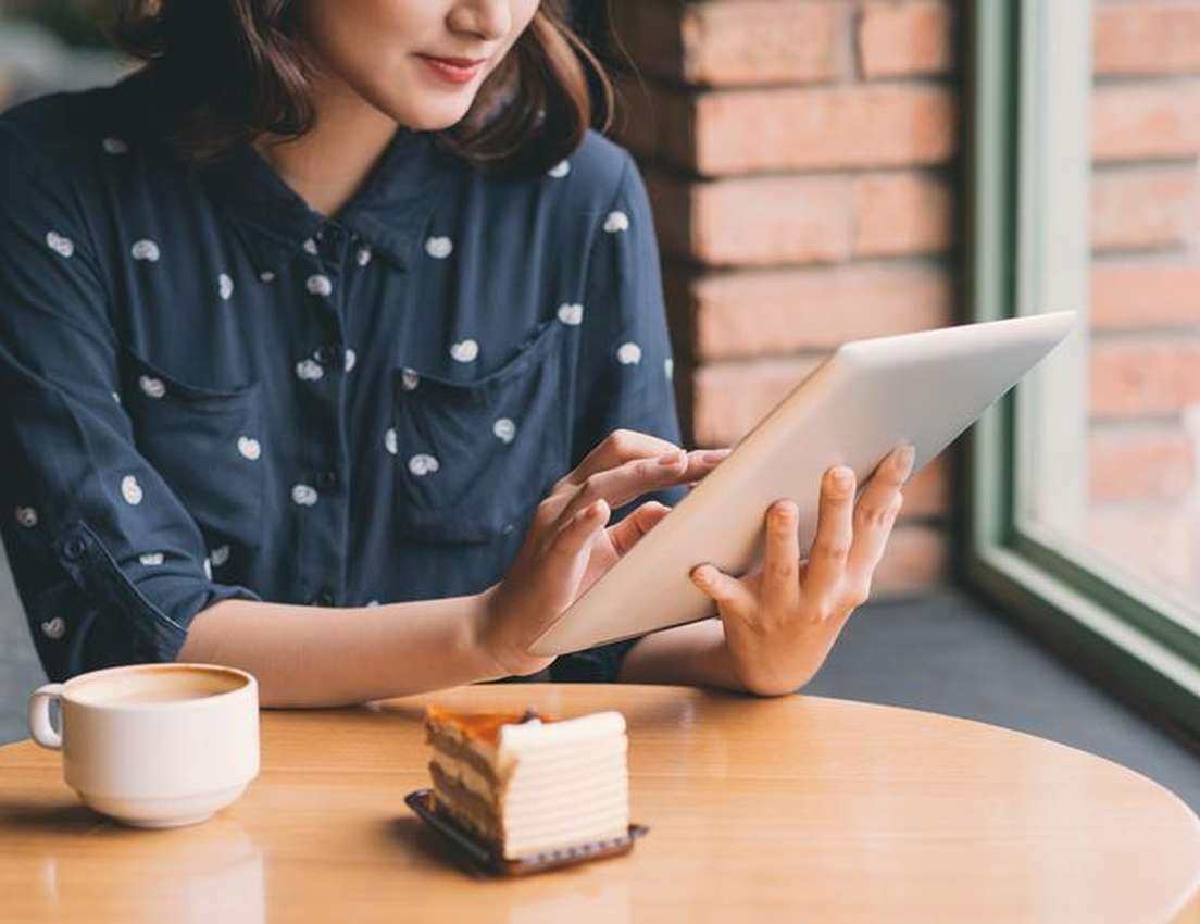 Woman wearing a dark polka dot shirt, browsing a tablet at a cafe table with a cup of coffee and a slice of cake.