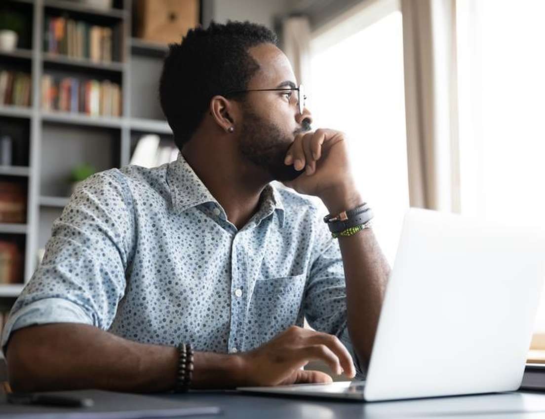 Man in a patterned shirt sits at a desk, gazing thoughtfully out a window, with a laptop open in front of him and bookshelves