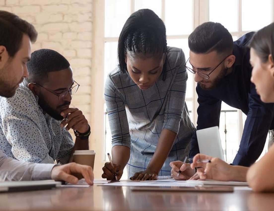 Diverse professionals huddle around a table, reviewing documents. A woman in the center writes on papers while others observe