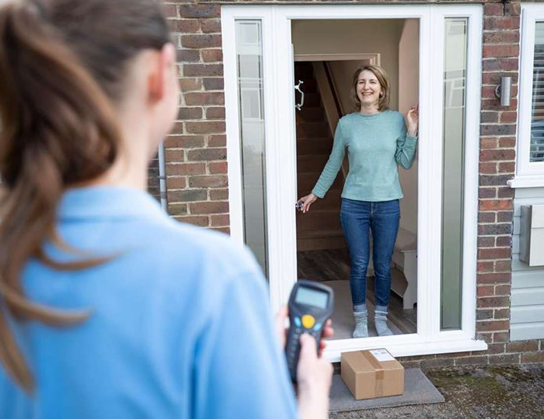 A delivery person, back to camera, holds a scanner while a smiling woman stands at her open front door. A package rests on th