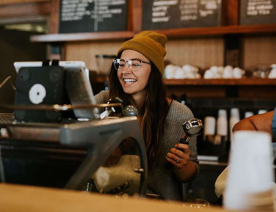 A smiling barista in a yellow beanie and glasses holds a portafilter behind a coffee bar, looking at a tablet display.