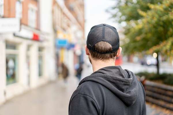 Back of a person in a black mesh cap and hoodie, looking down a blurry street with shops and trees on either side.