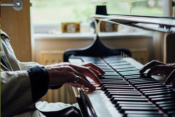 Aged hands with a ring play a black grand piano, fingers pressing keys. Soft light from a window illuminates the scene.