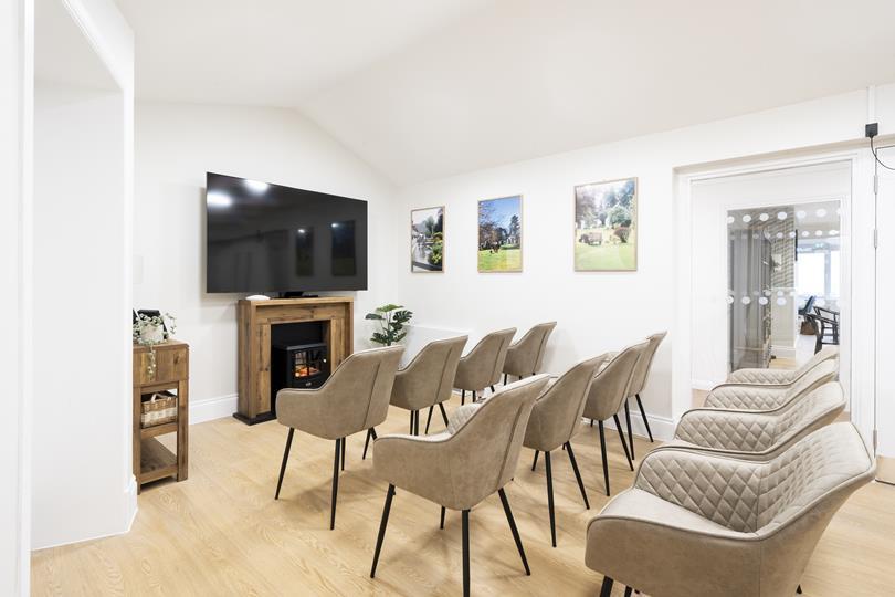 A bright, modern communal room with rows of beige chairs facing a TV above a wooden fireplace. Light wood floor, white walls