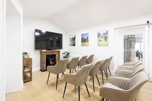A bright, modern communal room with rows of beige chairs facing a TV above a wooden fireplace. Light wood floor, white walls 