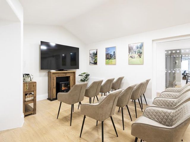A bright, modern communal room with rows of beige chairs facing a TV above a wooden fireplace. Light wood floor, white walls with artwork.