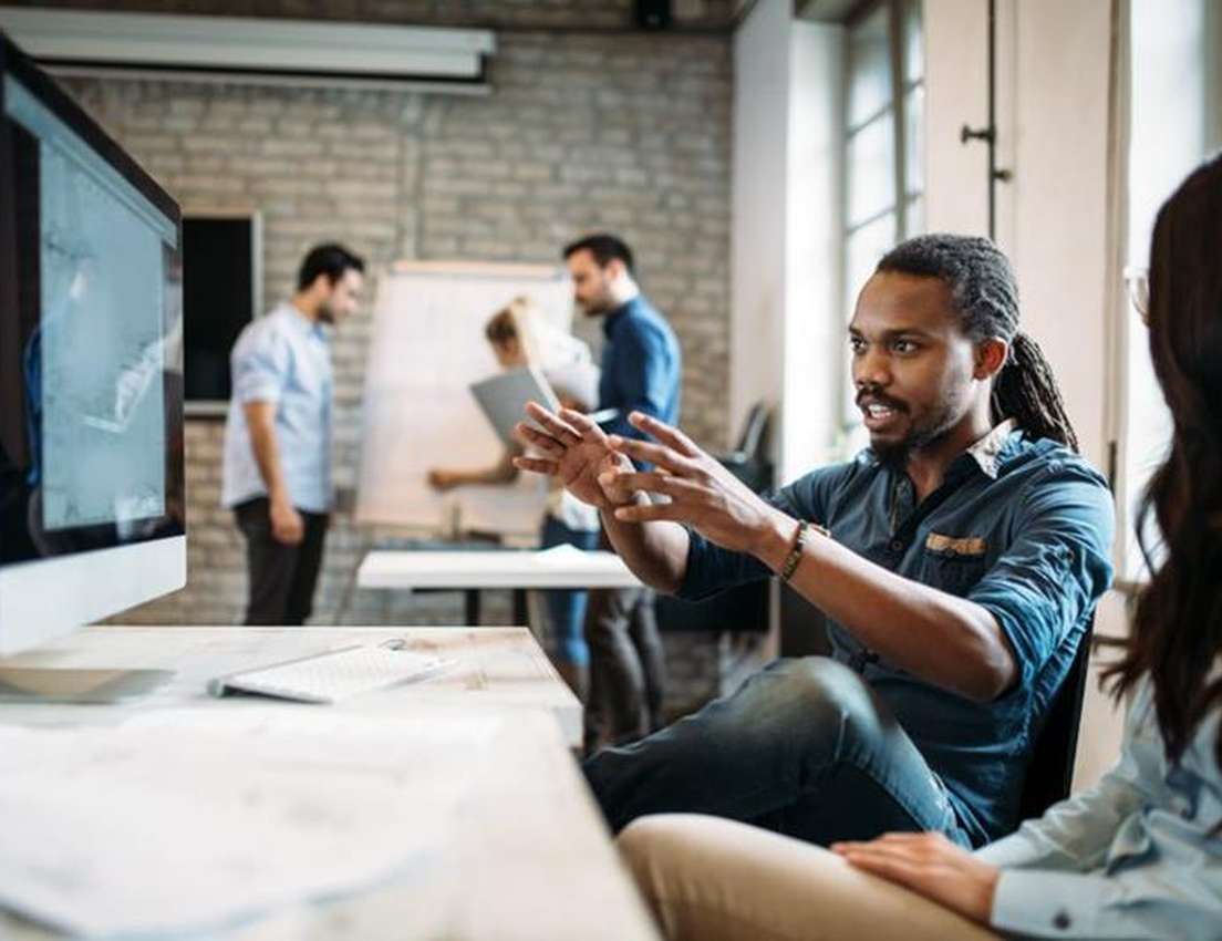A man with dreadlocks gestures while speaking to a woman in a modern office with computers and blurred people in the backgrou