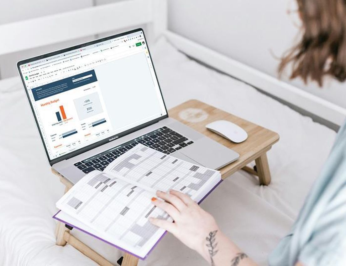 Person in bed manages finances using a laptop with a monthly budget spreadsheet and an open planner on a wooden bed tray.