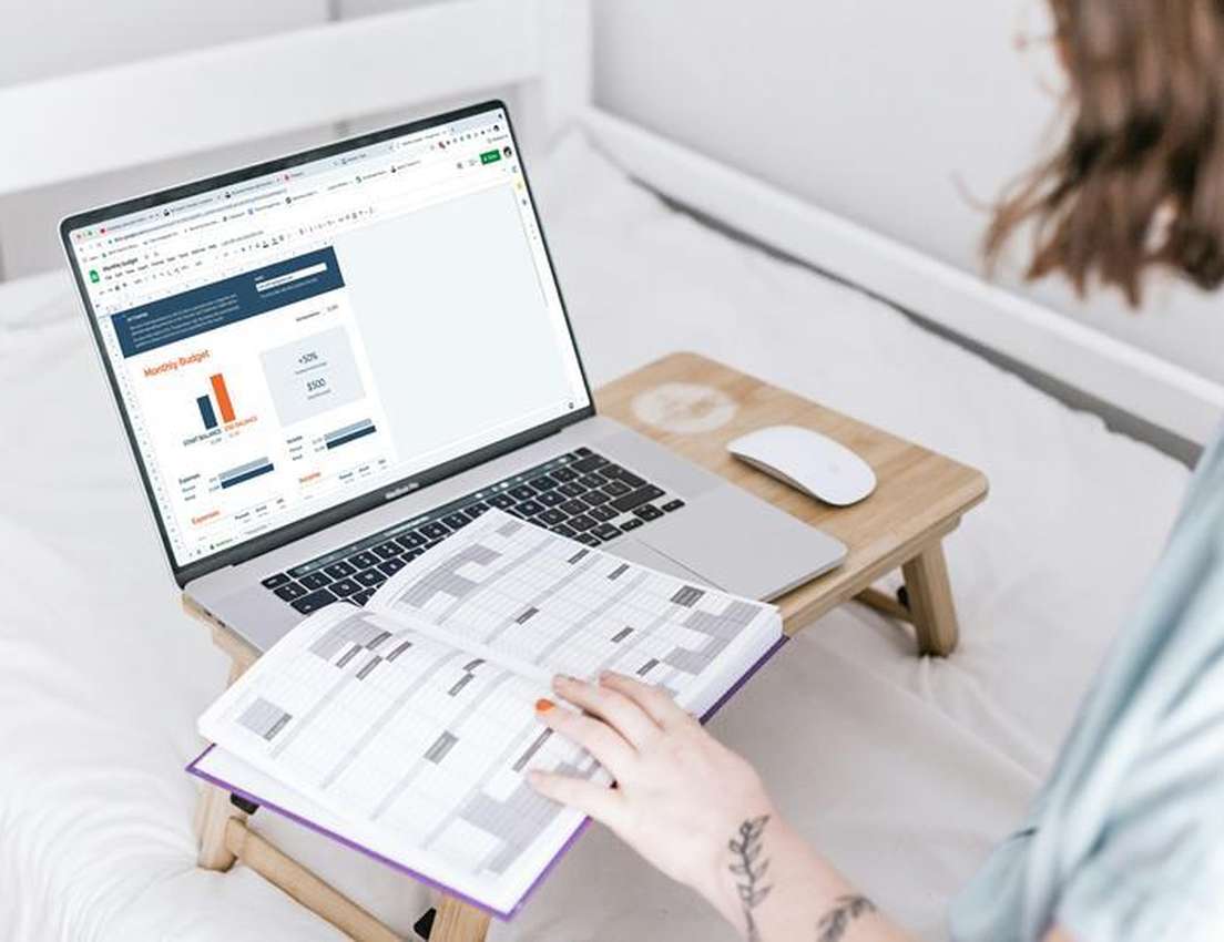 Person in bed manages finances using a laptop with a monthly budget spreadsheet and an open planner on a wooden bed tray.