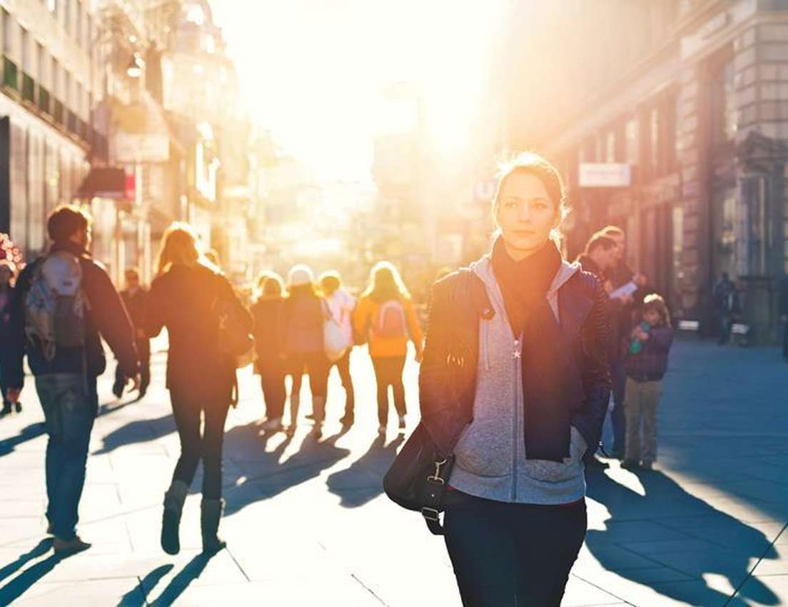 A woman in a jacket and scarf walks on a sun-drenched city street, surrounded by blurred people and long shadows.